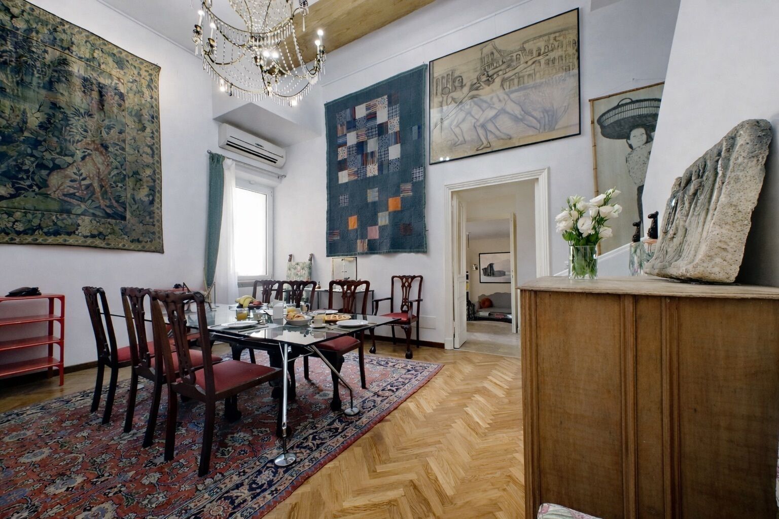 Een eetruimte met een grote tafel, stoelen en een kroonluchter. Er hangen kunstwerken aan de muur en een houten kast met een bloevas.