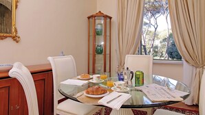 A dining area with a glass table, white chairs, a wooden sideboard, and a window with curtains. - Borghese Park View (Rome)