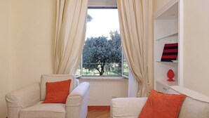 A room with two armchairs, a window with curtains, a shelf with a red object, and a view of trees outside. - Borghese Park View (Rome)