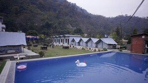 A swimming pool with a swan float, surrounded by a building with multiple rooms, a grassy area, and a hilly backdrop.