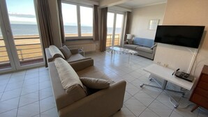 Un soggiorno spazioso con un divano grande, un tavolino da caffè e una TV a schermo piatto. Grandi finestre offrono vista sulla spiaggia.