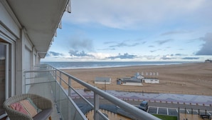 Un balcón con vista a una playa, una silla y un edificio pequeño con banderas.
