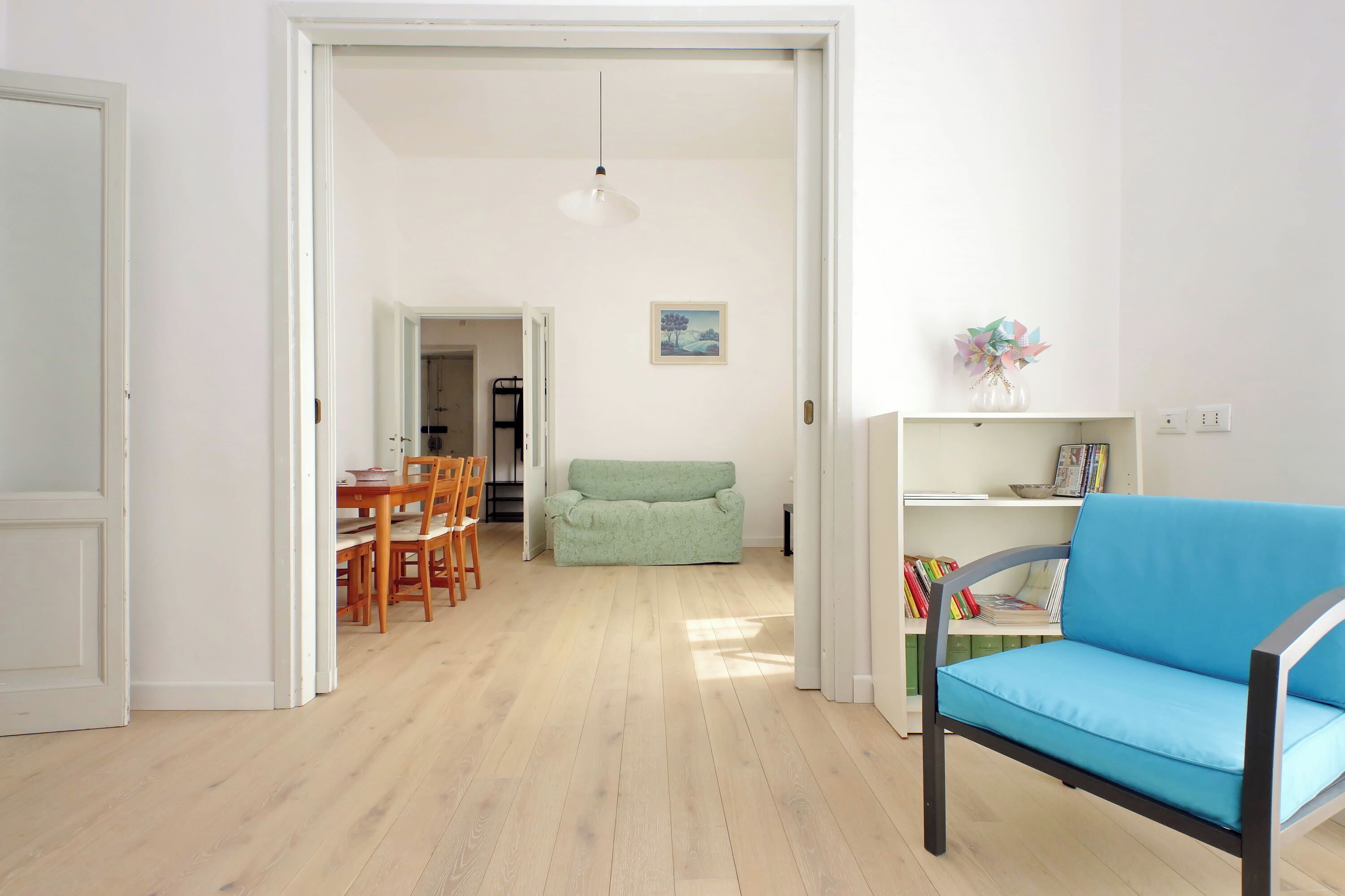 A spacious living area with a wooden floor, a green sofa, a blue armchair, and a bookshelf.