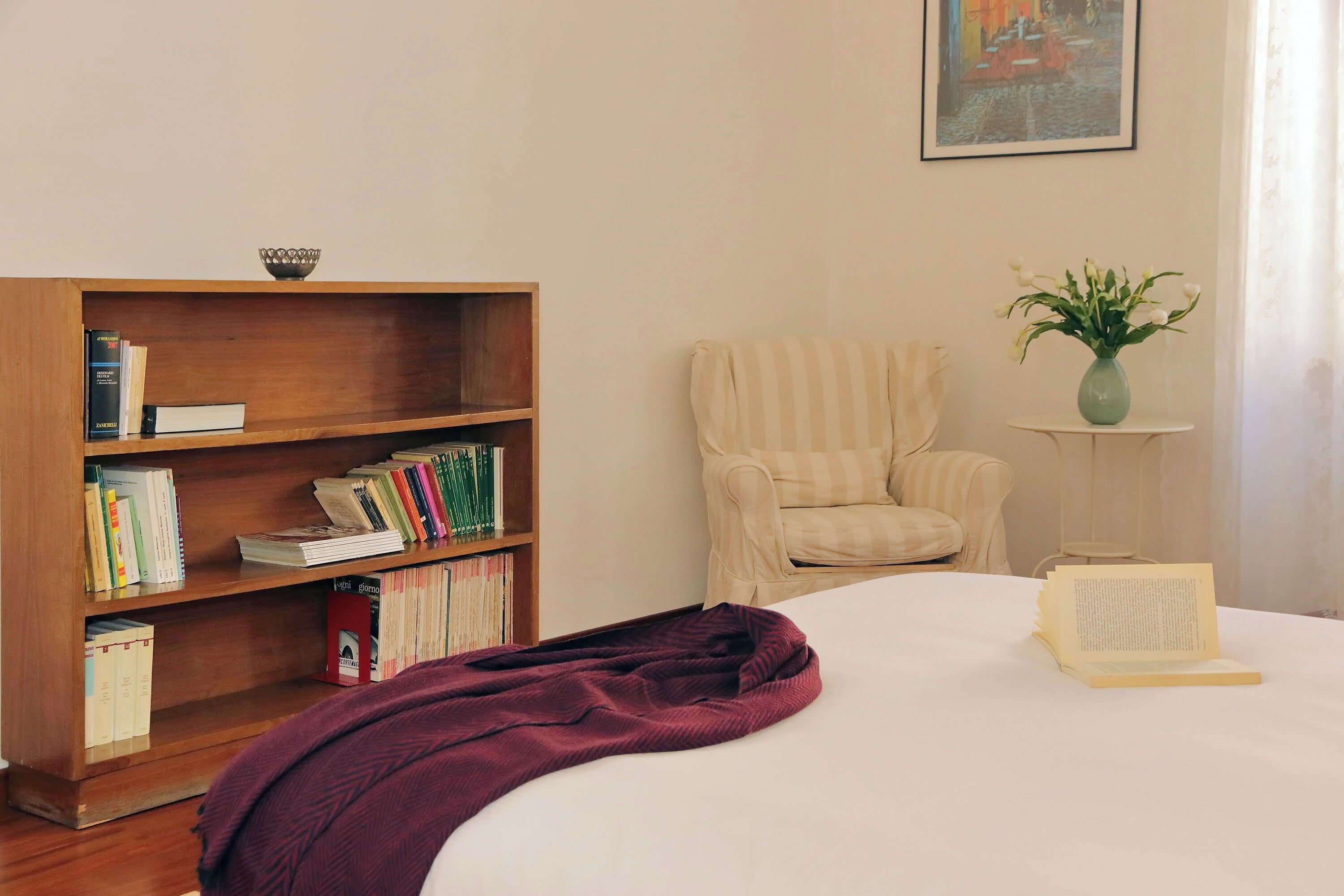 Una camera da letto con una libreria in legno, una poltrona a righe, un tavolino con un vaso e un letto con coperta viola.