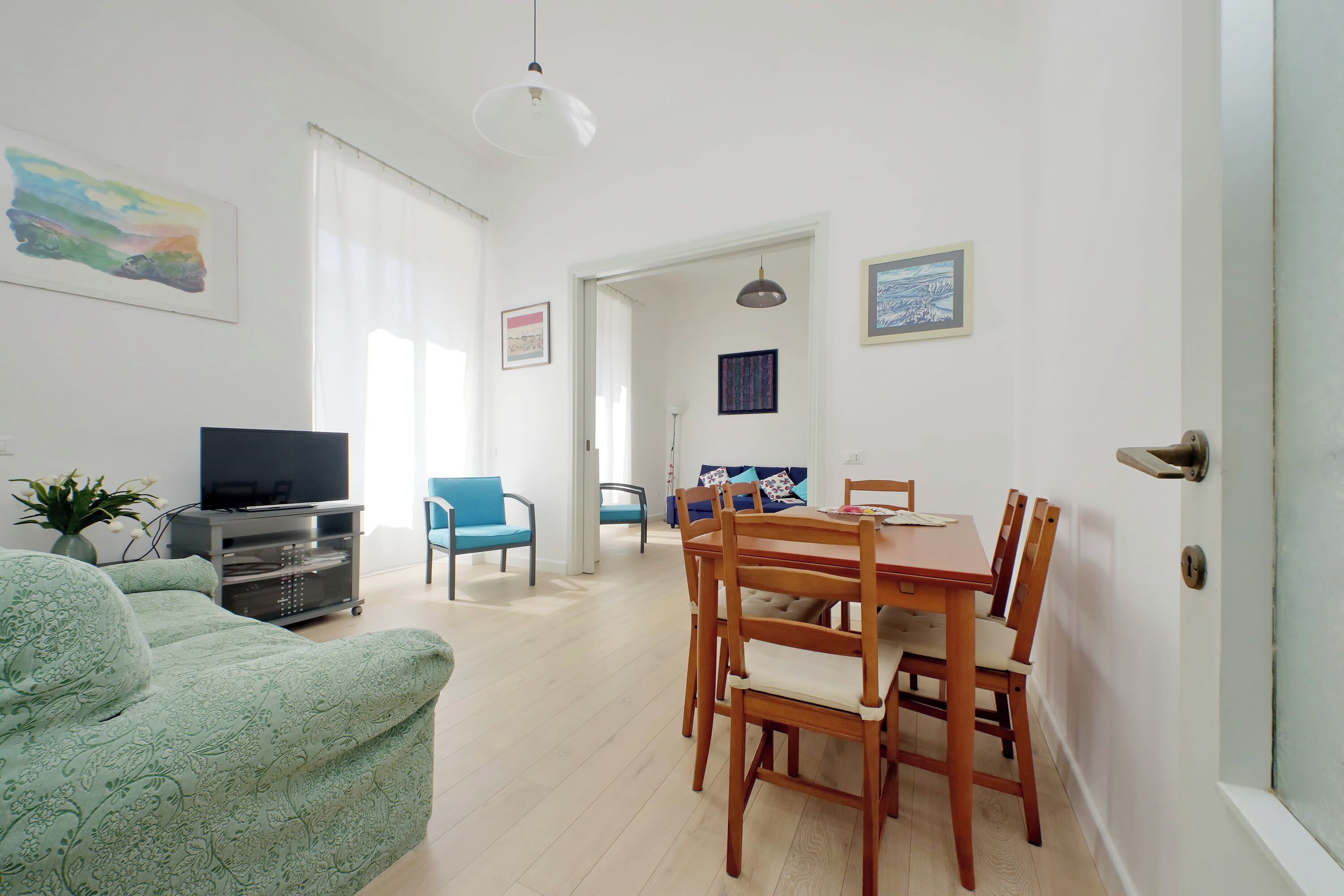 A living room with a sofa, TV, and dining table with chairs. There is a plant, a painting, and a door with a handle.