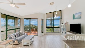 A spacious living room with a large sliding glass door leading to a balcony with a view of the ocean. There is a sofa, a coffee table, and a TV on a wooden stand.