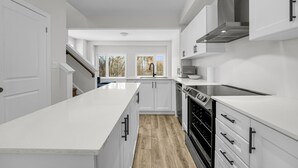 Une cuisine moderne avec des meubles blancs, un îlot central, des appareils en acier inoxydable et une vue sur les arbres grâce à de grandes fenêtres.