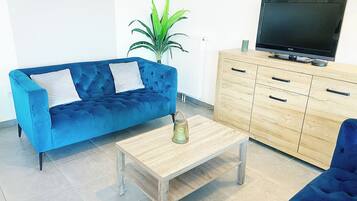 Ein Wohnzimmer mit blauem Sofa, einem Holztisch, einem Fernseher auf einer Holzkommode und einer Topfpflanze.