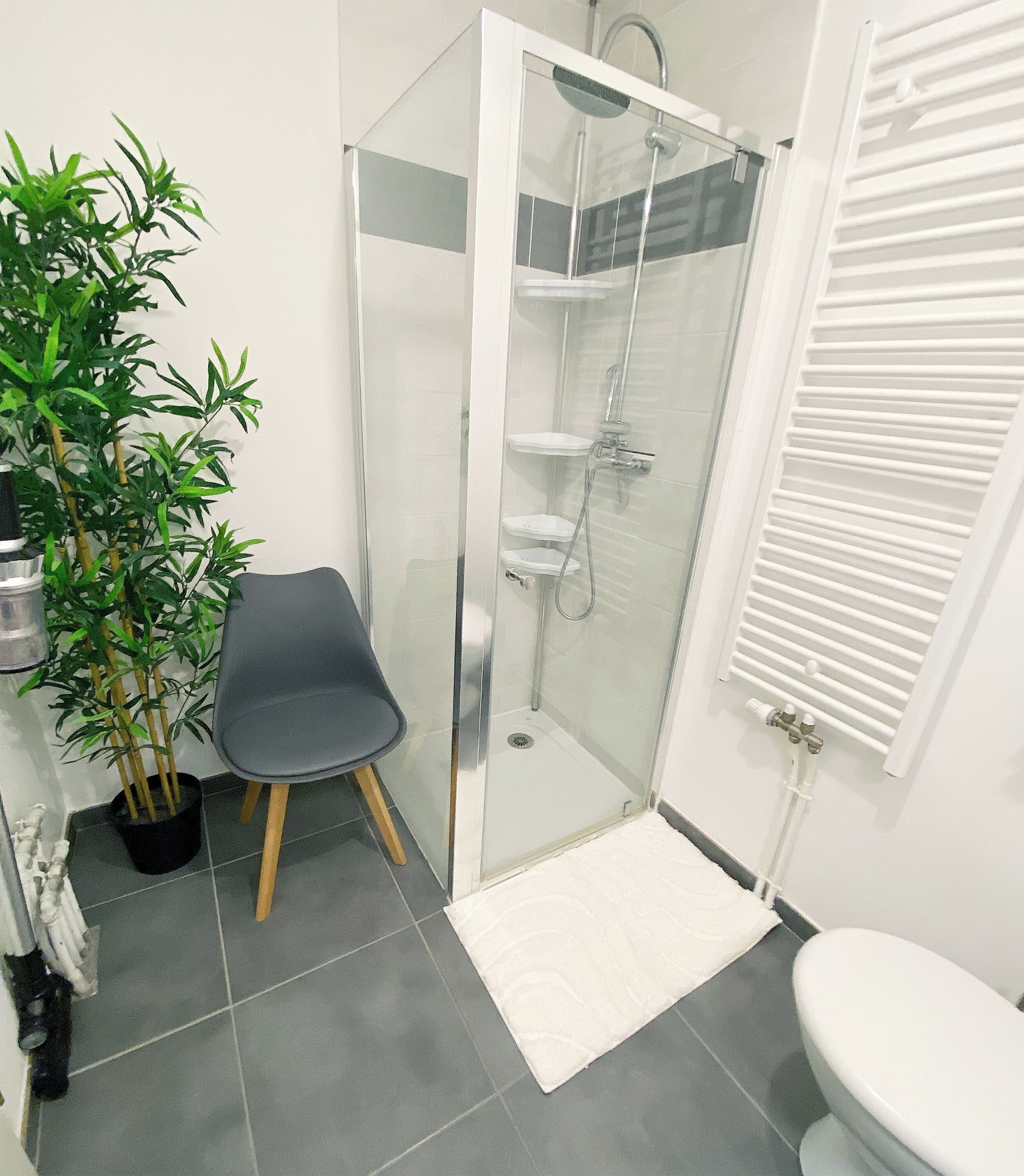 A bathroom with a glass-enclosed shower, a toilet, a sink, a potted plant, and a chair.