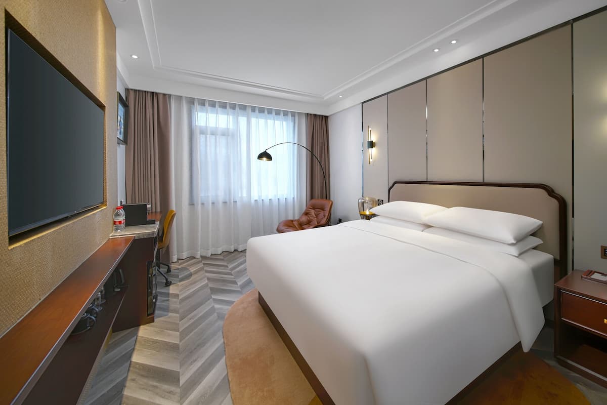 Habitación de hotel con una cama grande, un escritorio con televisor de pantalla plana, una silla, una lámpara de escritorio y un amplio ventanal con cortinas.