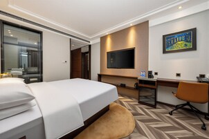 A hotel room with a bed, desk, chair, TV, and artwork on the wall.