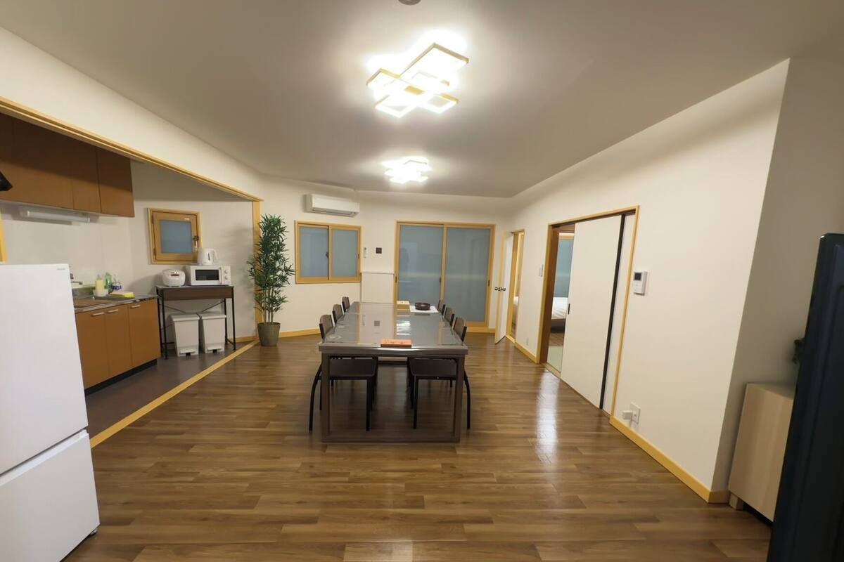 A spacious room with a wooden floor, a central dining table, a kitchen area with cabinets and appliances, and a potted plant.