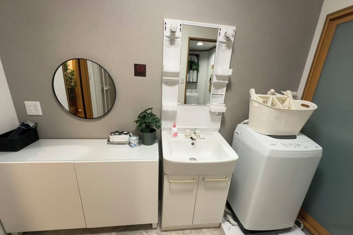 A bathroom with a white sink, a mirror, a small potted plant, a black bag, and a white laundry basket.