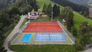 Una cancha de tenis rodeada por una cerca, con un edificio al fondo y un paisaje montañoso.