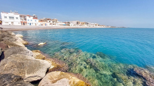 A coastal town with buildings along the waterfront, clear blue sea, and rocky shoreline.