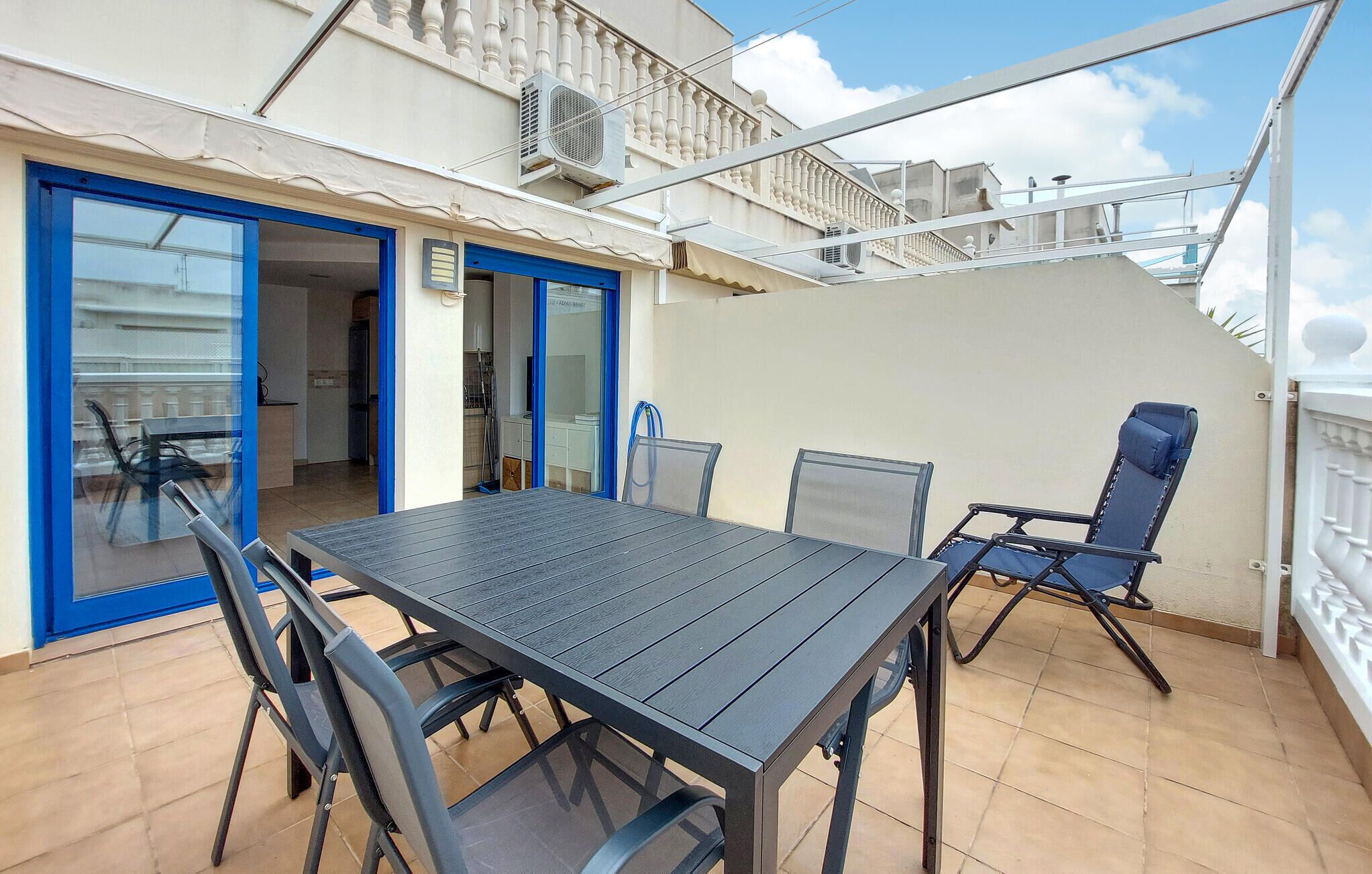 Une terrasse extérieure avec une table et des chaises, une porte bleue et un escalier menant à un niveau supérieur.