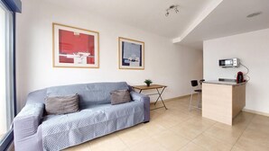 A compact living space with a sofa, a small table with a plant, a kitchenette with a microwave, and two abstract paintings on the wall.