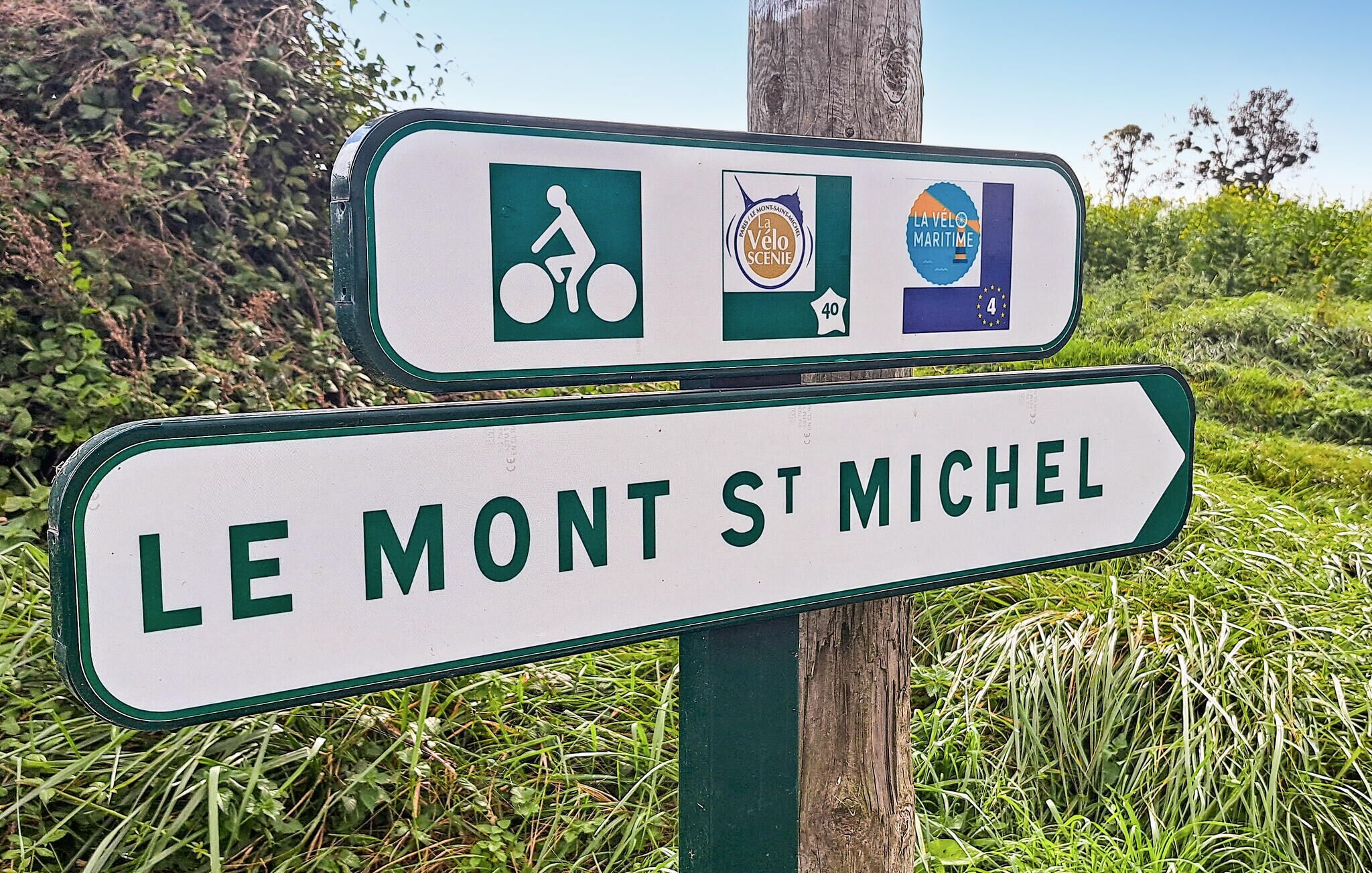 Un panneau de signalisation indiquant la direction du Mont-Saint-Michel, avec des symboles représentant un itinéraire cyclable, un site pittoresque et un thème maritime.