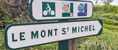 Un panneau de signalisation indiquant la direction du Mont-Saint-Michel, avec des symboles représentant un itinéraire cyclable, un site pittoresque et un thème maritime.