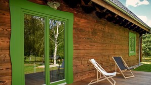 A wooden cabin with green-framed glass doors, a deck with two lounge chairs, and a view of trees and a gazebo. - Cozy home in Czaplinek with lake view (Czaplinek)