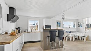 Une cuisine moderne avec des armoires blanches, un comptoir en bois et un îlot central doté de tabourets de bar. On y trouve également un coin repas avec une table et des chaises blanches, et une vue sur l’extérieur grâce à de grandes fenêtres.