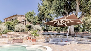 Un área de piscina con suelo de piedra, una zona de estar con cojines y un parasol grande.