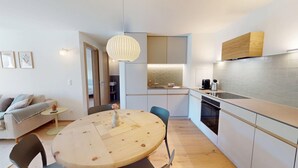 A compact living space with a round wooden table, a sofa, a kitchen with a modern design, and a pendant light.