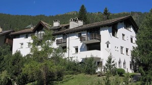 A multi-story building with balconies, surrounded by greenery and set against a mountainous backdrop.