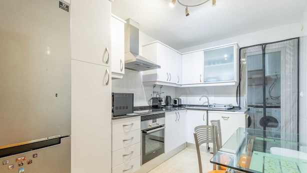 A modern kitchen with white cabinets, a built-in oven, a microwave, and a glass dining table with orange chairs. - Diana Apartment (Teulada)