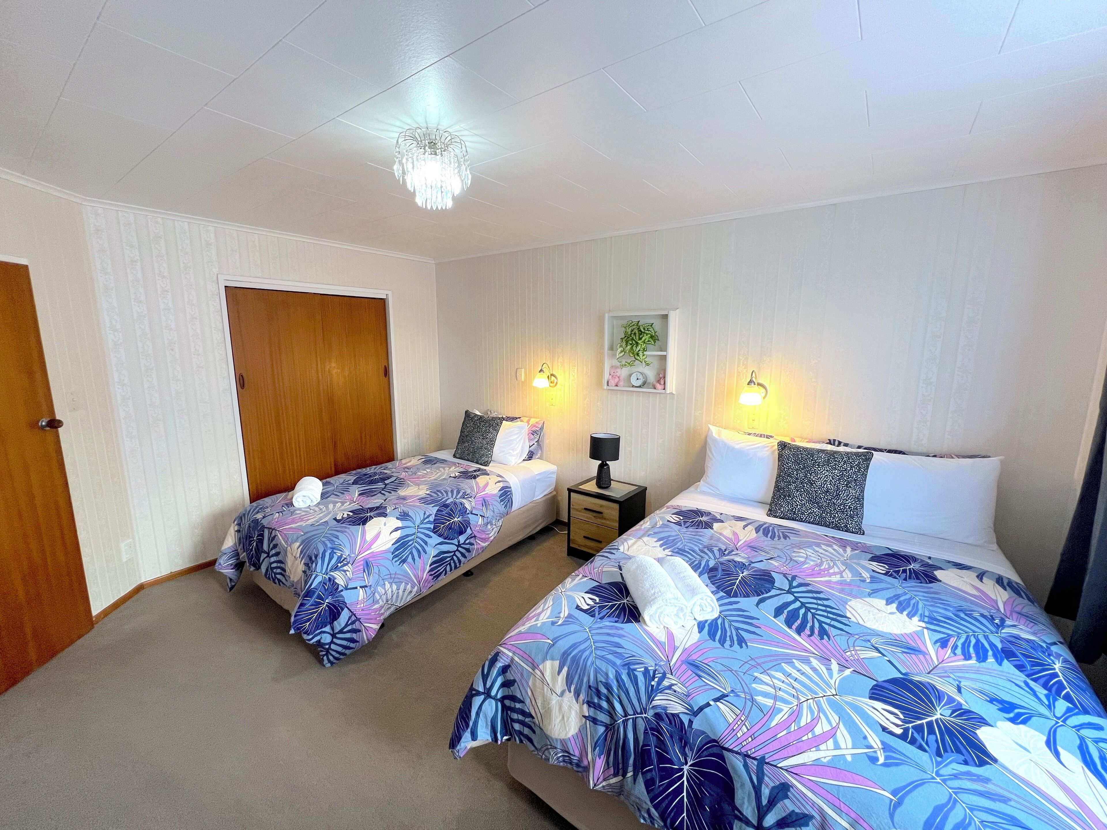 Una habitación con dos camas, cada una con cubrecama floral, una puerta de madera, una mesita de noche con lámpara y luces en la pared.