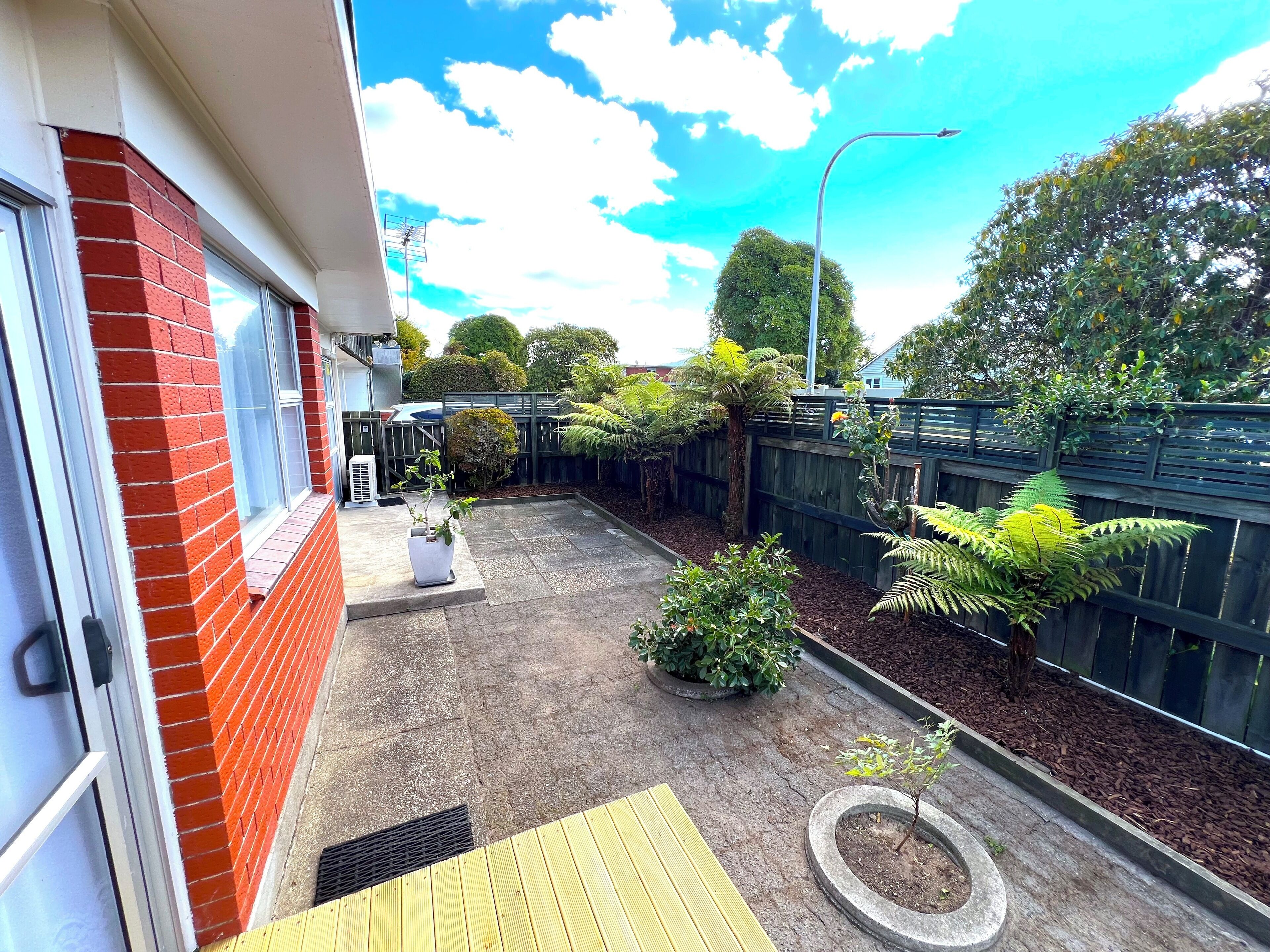 Un jardin à l’arrière d’une maison d’habitation, avec un mur en briques, une terrasse en bois, un petit jardin planté de différentes espèces végétales et une clôture.