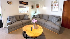 A living room with a grey sectional sofa, a round wooden coffee table with a vase of flowers, a clock on the wall, and a shelf with decorative items. - Rotorua Central Accommodation  (Rotorua)