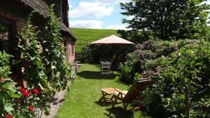 A garden with a table and chairs, a thatched-roof building, and a red umbrella. - Idyllic thatched house directly on the Eider dike for 2 to 10 people (Oldenswort)