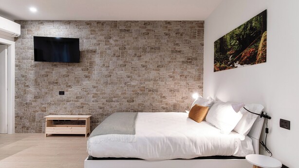 A modern bedroom with a stone wall, a mounted TV, a bed with white bedding, a small wooden nightstand, and a painting of a forest scene. - Victoria Studio 1  (Como)