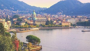 A riverside cityscape with historic buildings, a prominent church, and a tree-lined promenade. - Victoria Studio 1  (Como)