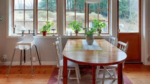 A dining area with a wooden table and chairs, large windows with plants, and a view of greenery outside.