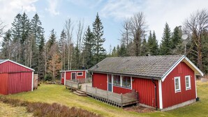 A red wooden cabin with a porch and a smaller adjacent shed, surrounded by a forested area.