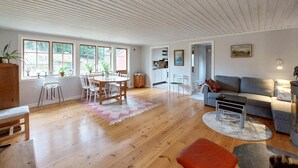 A spacious living area with a wooden floor, a dining table with chairs, a grey sofa, and a small orange stool.