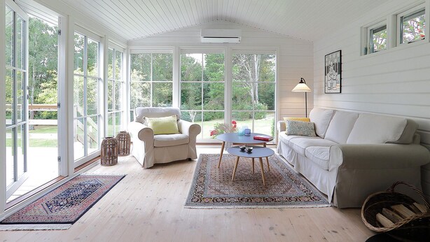 Ein geräumiges Zimmer mit großem Fenster, weißer Decke, beiger Couch, weißem Sessel, kleinem Tisch und einer Stehlampe.