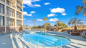Una piscina con tumbonas y palmeras, rodeada por un edificio con balcones.
