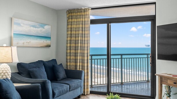 Una habitación con sofá, un cuadro, cortina, televisión y vista a la playa.
