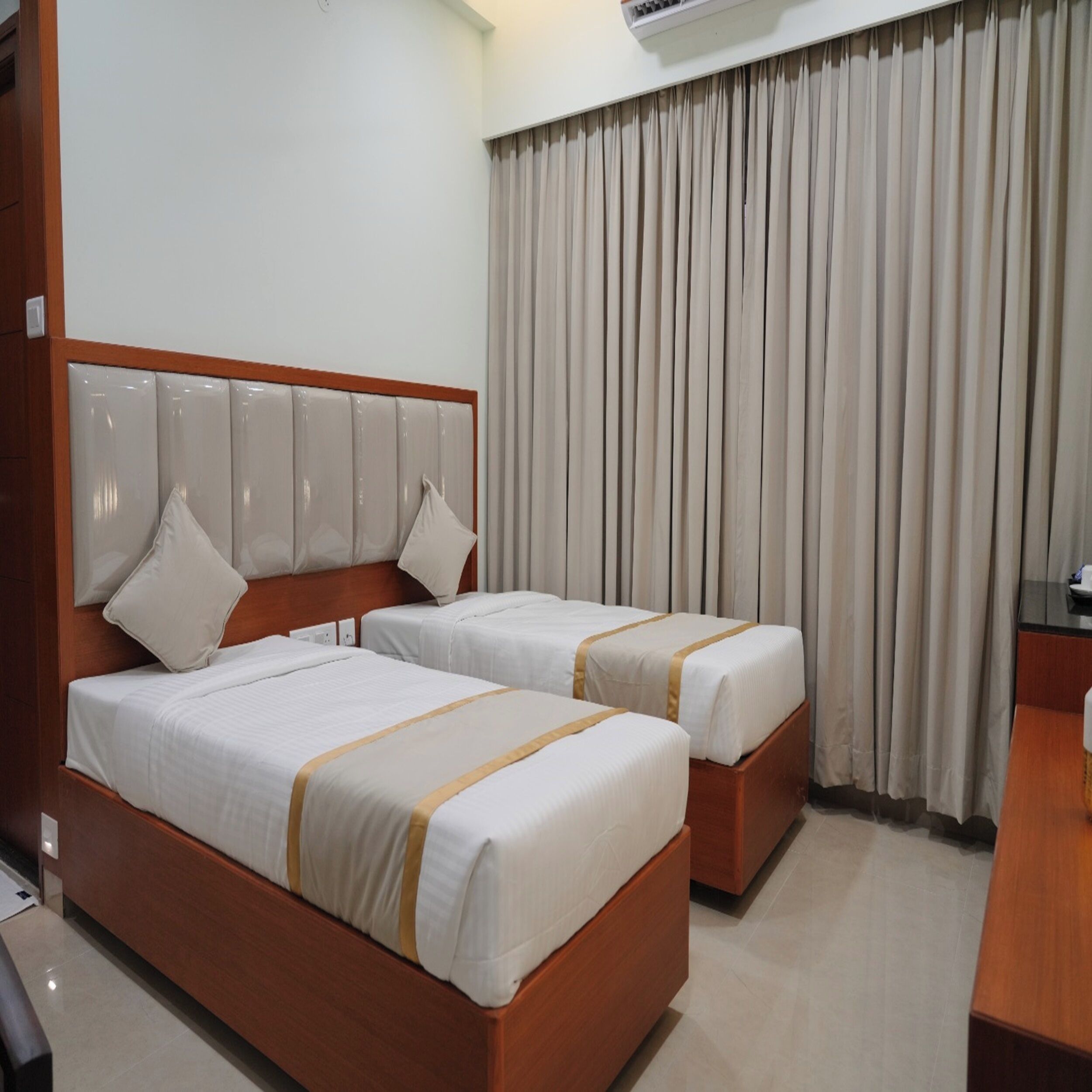 Ein Hotelzimmer mit zwei Betten, einem hölzernen Kopfteil, einem großen Fenster mit Vorhängen und einem Schreibtisch mit Stuhl.