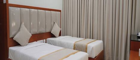 Ein Hotelzimmer mit zwei Betten, einem hölzernen Kopfteil, einem großen Fenster mit Vorhängen und einem Schreibtisch mit Stuhl.