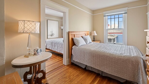 A bedroom with a large bed, a bedside table, a lamp, and a window with a view of buildings. - Downtown Luray Historic Home: Shenandoah • Caverns (Luray)