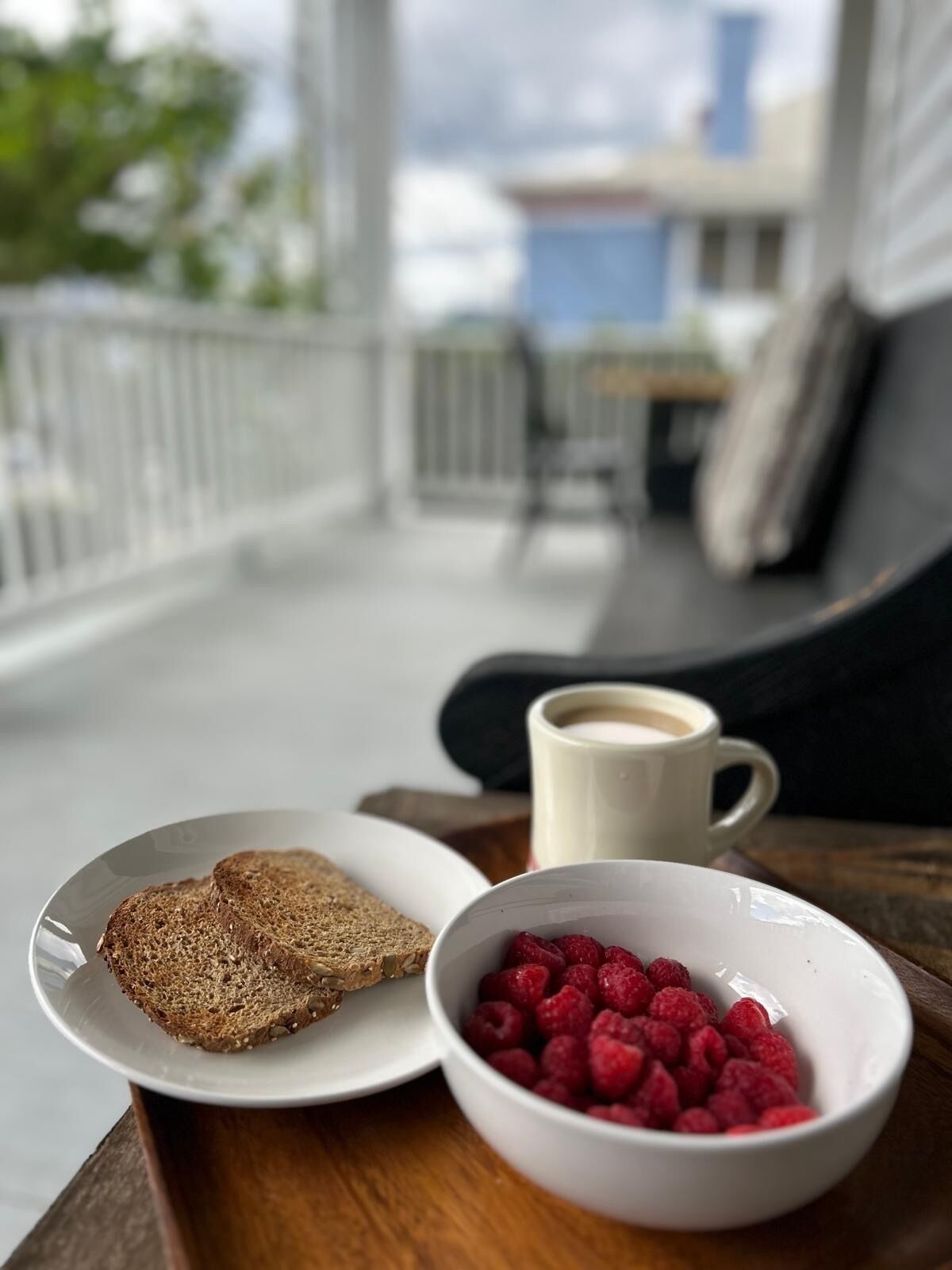 Ett frukostbricka med rågbröd, en skål med hallon och en kopp kaffe står på ett träbord i ett rum med utsikt över byggnader och grönområden.