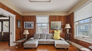 A living room with a sectional sofa, wooden paneled walls, and two windows with blinds. - Downtown Luray Historic Home: Shenandoah • Caverns (Luray)