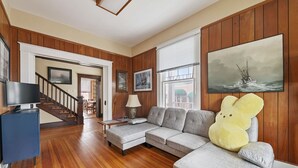 A living room with a wooden floor, a grey sofa, a television, and a large painting of a ship on the wall. - Downtown Luray Historic Home: Shenandoah • Caverns (Luray)