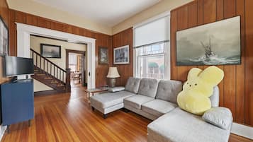 A living room with a wooden floor, a grey sofa, a television, and a large painting of a ship on the wall.