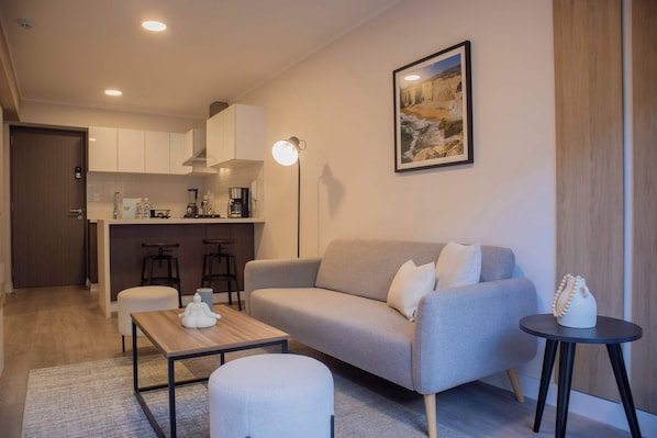 A modern living room with a sofa, ottoman, coffee table, and a small side table. A kitchen area is visible in the background with bar stools.
