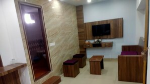 A modern living room with a wooden floor, a flat-screen TV mounted on the wall, a small wooden table, and a bench. - Hotel Tak (Udaipur)